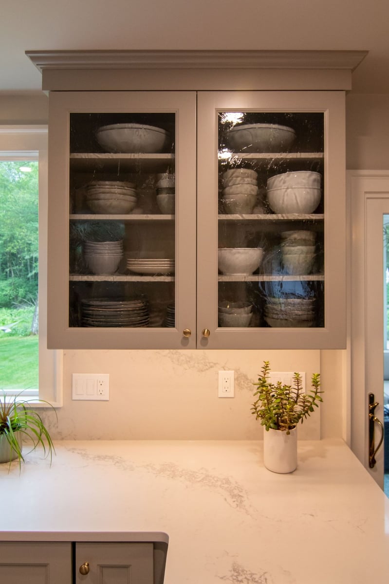 Glass-front cabinet with stacked neutral dishware above a white quartz counter and small potted plants.